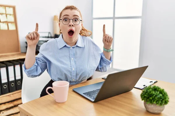 Ofiste çalışan genç kızıl saçlı kadın bilgisayarını kullanıyor. Şaşırmış, şaşırmış ve parmaklarıyla işaret ediyor ve kollarını kaldırıyor.. 