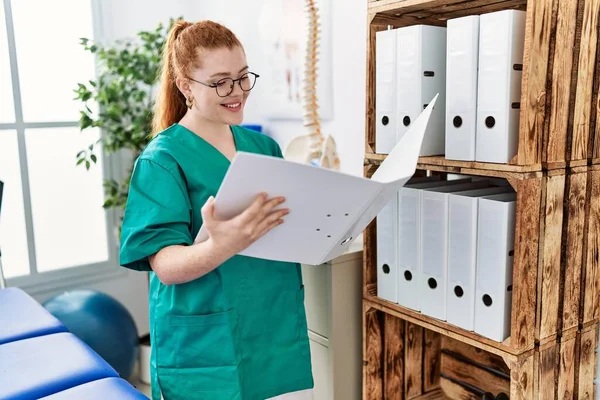 Fsiolog üniforması giyen genç kızıl saçlı kadın klinikte klasör tutuyor.