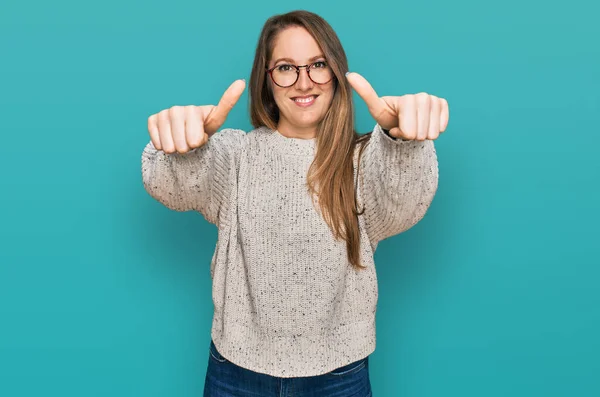 Young blonde woman wearing casual sweater and glasses approving doing positive gesture with hand, thumbs up smiling and happy for success. winner gesture.