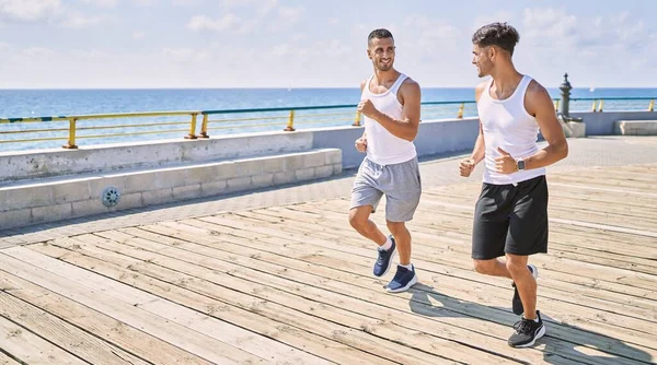 İki İspanyol erkek, sportif, gülümseyen bir çift, deniz kenarında koşuyorlar.