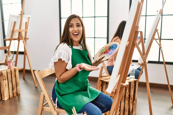 若いヒスパニック系のアーティストの女性は コピースペースを示す手を開いて手のひらで脇を指しているアートスタジオでキャンバス上に絵を描く 広告を提示笑顔興奮幸せ — ストック写真
