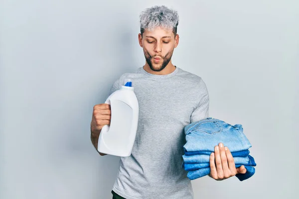 Young hispanic man with modern dyed hair holding jeans for laundry and detergent bottle making fish face with mouth and squinting eyes, crazy and comical.