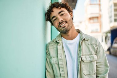 Yakışıklı, sakallı, güneşli bir günde mutlu bir şekilde gülümseyen bir İspanyol. Duvara yaslanmış.