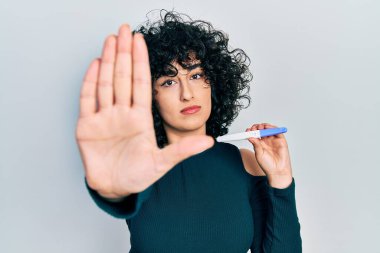 Orta Doğulu genç bir kadının elinde hamilelik testi sonucu açık elle dur işareti yaparak ciddi ve kendinden emin bir ifadeyle savunma hareketi yapması. 