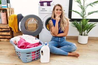 Çamaşır sepetinin yanında oturan genç ve güzel bir kadın kameraya kollarını kavuşturup gülümseyerek ve çenesini kaldırarak bakıyor. Olumlu düşünmek. 