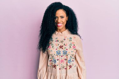 Orta yaşlı Afro-Amerikalı kadın günlük kıyafetler giyiyor. Pozitif ve mutlu görünüyor. Kendine güvenen bir gülümsemeyle dişlerini gösteriyor. 