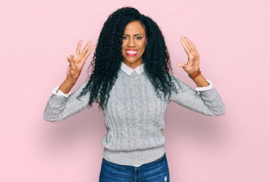 Orta yaşlı Afro-Amerikalı kadın gündelik giysiler giyip 8 numaralı parmaklarıyla işaret ederken kendinden emin ve mutlu gülümsüyor.. 