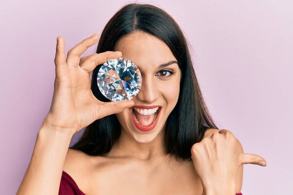 Young hispanic woman holding brilliant diamond stone on eye pointing thumb up to the side smiling happy with open mouth 