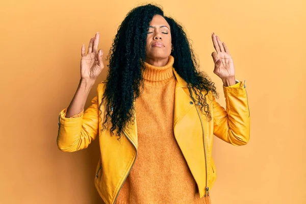 Orta yaşlı Afro-Amerikalı kadın yün kazak ve deri ceket giyiyor gözleri kapalı gülümsüyor parmaklarıyla meditasyon hareketi yapıyor. yoga konsepti. 