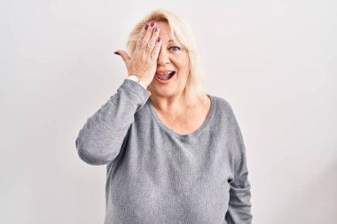 Beyaz arka planda duran orta yaşlı beyaz bir kadın bir gözünü eliyle kapatıyor, yüzünde kendine güvenen bir gülümseme ve şaşırtıcı bir duygu var.. 