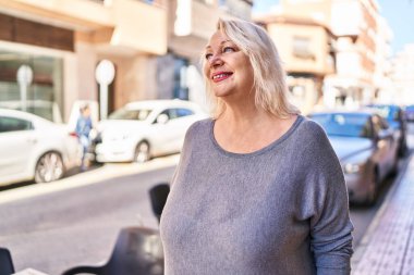 Orta yaşlı sarışın kadın sokakta kendinden emin bir şekilde gülümsüyor.