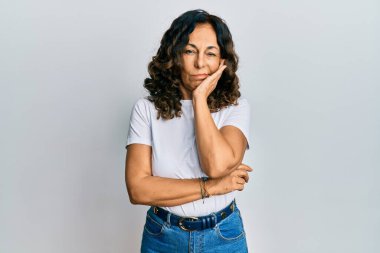 Orta yaşlı, beyaz tişört giyen İspanyol bir kadın kollarını kavuşturarak depresyondan sıkılmış ve yorgun görünüyor.. 