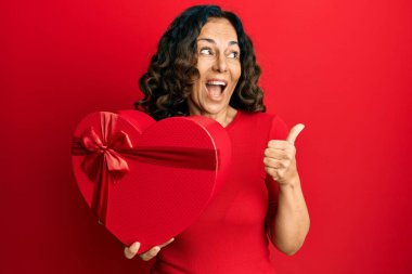 Orta yaşlı İspanyol bir kadın sevgililer günü hediyesi tutuyor. Parmağını kaldırıp gülümsüyor ve ağzı açık gülümsüyor. 