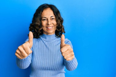 Orta yaşlı İspanyol kadın gündelik giysiler giyip olumlu jestler yapıyor, gülümsüyor ve başarı için seviniyor. kazanan jest. 