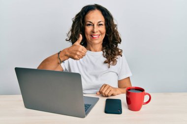 Ofiste çalışan orta yaşlı güzel bir kadın bilgisayarında mutlu ve olumlu gülümseyen bir dizüstü bilgisayar kullanıyor, başparmağı mükemmel ve onay işareti yapıyor. 