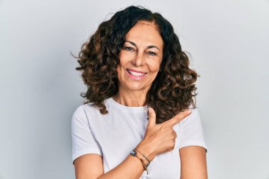 Orta yaşlı İspanyol kadın, günlük beyaz tişört giyiyor. Gülümsüyor, eli ve parmağı yana doğru uzanıyor. 