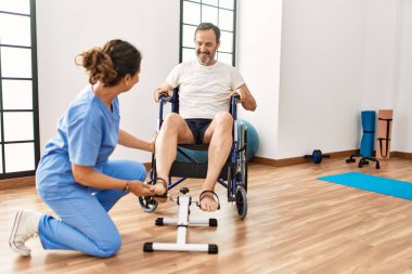 Orta yaşlı bir adam ve kadın fizik tedavi kliniğinde tekerlekli sandalyede pedal çeviriyorlar.