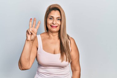 Orta yaşlı İspanyol kadın kolsuz gömlekli rahat giyiniyor ve üç numaralı parmaklarını gösterip gülümsüyor ve mutlu.. 