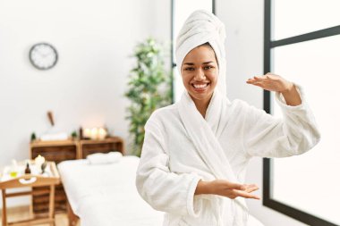 Havlu ve bornoz giymiş genç esmer kadın güzellik merkezinde el kol hareketleri yaparak büyük ve büyük bir işaret, ölçü sembolü gösteriyor. Kameraya bakıp gülümsüyor. ölçüm kavramı. 