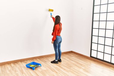 Arkadaki genç Latin kadın boş odada silindir kullanarak duvarları boyuyor.
