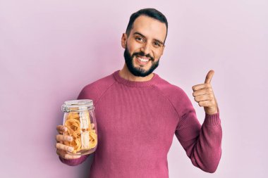 Elinde makarna dolu sakallı genç adam mutlu ve pozitif gülümsüyor, başparmağıyla mükemmel ve onay işareti yapıyor. 