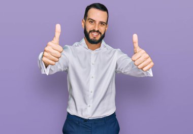 Sakallı, iş gömleği giyen, el hareketlerini onaylayan, gülümseyen ve başarı için sevinen genç bir adam. kazanan jest. 