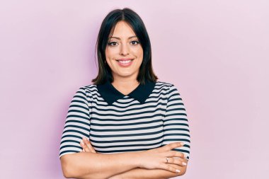 Gündelik kıyafetler giyen, kollarını kavuşturup kameraya gülümseyen genç İspanyol kadın. pozitif kişi. 