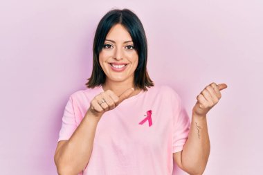 Tişörtüne pembe kanser kurdelesi takmış genç İspanyol bir kadın. Arka tarafı işaret ediyor. El ve baş parmakları havada, kendinden emin gülümsüyor. 