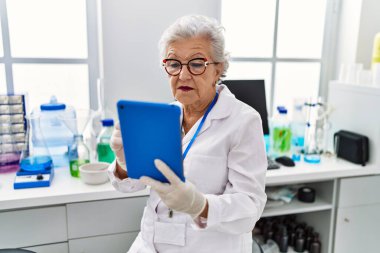 Laboratuvarda dokunmatik ped kullanan yaşlı gri saçlı kadın bilim adamı üniforması giyiyor.