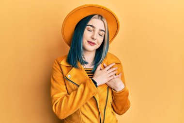 Sarı şapka ve deri ceket giyen, elleri göğsünde, gözleri kapalı gülümseyen ve yüzünde minnet ifadesi olan genç modern bir kız. sağlık kavramı. 