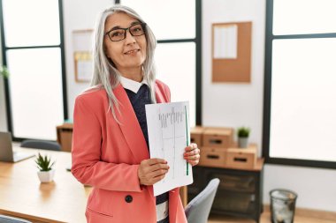 Orta yaşlı, kır saçlı bir iş kadını elinde belgelerle ofiste duruyor.