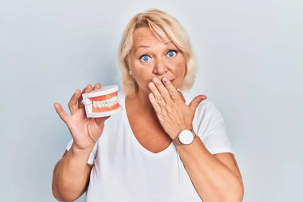 Middle Age Blonde Woman Holding Denture Covering Mouth Hand Shocked — Stock Photo, Image