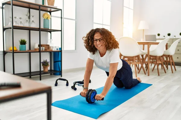 Orta yaşlı İspanyol kadın gülümsüyor. Karın kaslarını çalıştırırken evde tekerlekli sandalye kullanıyor.