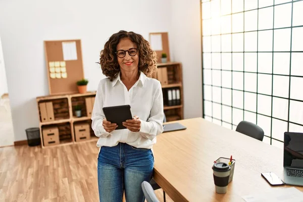 Orta yaşlı İspanyol kadın, ofiste dokunmatik ped kullanarak çalışırken kendine güvenerek gülümsüyor.