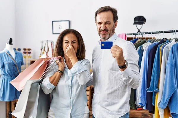 Hispanic middle age couple holding shopping bags and credit card smelling something stinky and disgusting, intolerable smell, holding breath with fingers on nose. bad smell