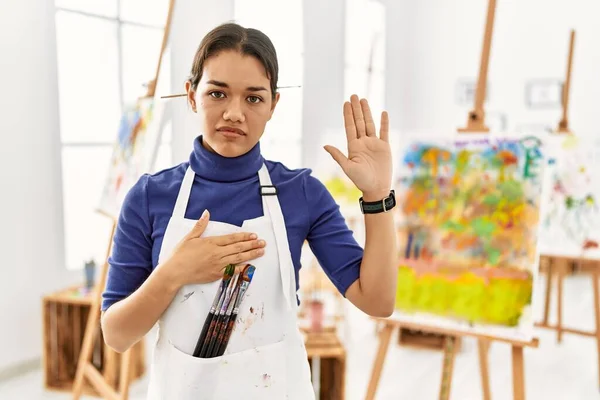 Young brunette woman at art studio swearing with hand on chest and open palm, making a loyalty promise oath