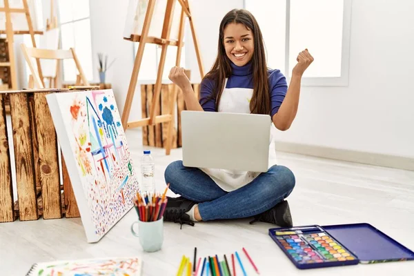年轻的黑发女子坐在艺术工作室的地板上 带着笔记本电脑 自豪地尖叫着 高举双臂庆祝胜利和成功 — 图库照片