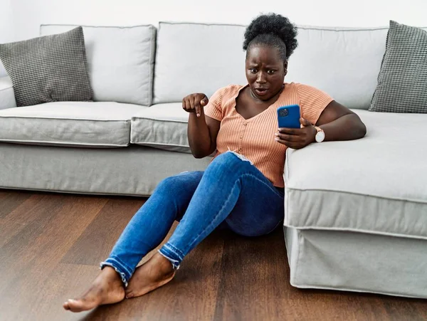 Young African Woman Using Smartphone Sitting Floor Home Pointing Fingers — Stock fotografie