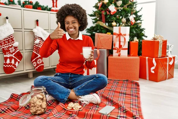 若いですアフリカ系アメリカ人女性とともにAfro毛食べるペストリーによって座ってクリスマスツリー笑顔と正 親指アップ行う優れたと承認サイン — ストック写真