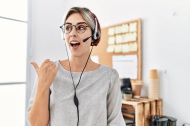 Young beautiful woman working at the office wearing operator headset pointing thumb up to the side smiling happy with open mouth 