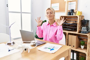 Ofiste çalışan beyaz bir kadın rahatlayıp gülümsüyor ve gözleri kapalı parmaklarıyla meditasyon yapıyor. yoga konsepti. 