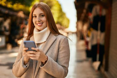 Genç kızıl saçlı kız şehirde akıllı telefon kullanarak mutlu bir şekilde gülümsüyor..