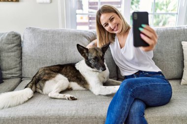 Genç beyaz kız kanepede oturmuş mutlu bir şekilde gülümsüyor köpekle evde akıllı telefonun yanında selfie çekiyor..