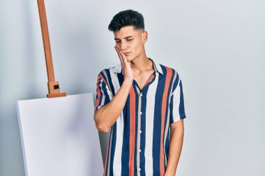 Dişlerinin ağrısından ya da diş ağrısından dolayı boş tuvalin yanında duran genç bir İspanyol acı dolu bir ifadeyle ağzına dokunuyor. dişçi 