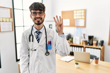 Ofiste doktor üniforması giyen ve steteskop takan İspanyol bir adam dört numarayı gösterip işaret ederken kendinden emin ve mutlu bir şekilde gülümsüyor.. 