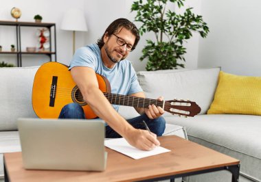 Orta yaşlı beyaz bir adam evde gitar ve dizüstü bilgisayar kullanarak şarkı besteliyor.