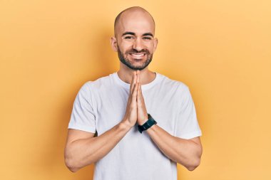 Gündelik beyaz tişört giyen genç kel adam ellerinin birarada dua ederek af diliyor kendinden emin bir şekilde gülümseyerek.. 