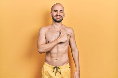 Yüzünde mutlu ve doğal bir ifade olan, elleri ve parmaklarıyla işaret eden, neşeli, mayo giymiş genç kel bir adam. 