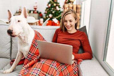 Genç Kafkasyalı kadın evde köpekle otururken dizüstü bilgisayar kullanıyor.