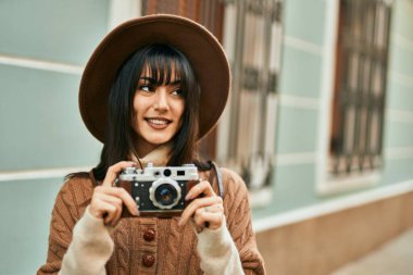 Kışlık şapkalı esmer kadın şehrin dışında klasik kamera kullanarak gülümsüyor.